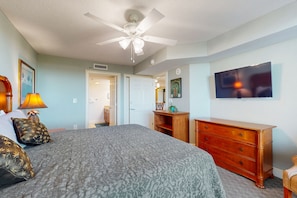 A bedroom with a bed, a dresser, a ceiling fan, a television, and a door leading to another room.