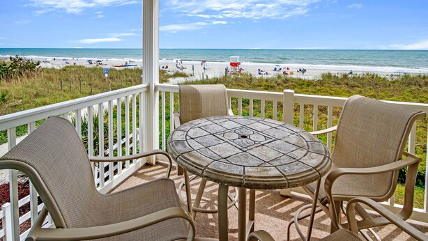 Een overdekt terras met een ronde tafel en stoelen, uitkijkend op een strand met mensen en parasols.