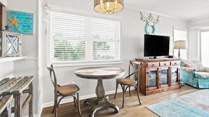 Een gezellige woonkamer met een ronde houten tafel, twee stoelen, een televisie op een houten kast en een groot raam met zonwering.