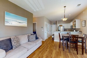 A modern living room with a beige sofa, patterned cushions, and a large landscape painting. A wooden dining table with chairs is in the center. The kitchen area is visible with white cabinets and a refrigerator.