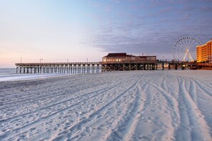 A beach with a pier extending into the water, a building on the pier, and a Ferris wheel in the background.