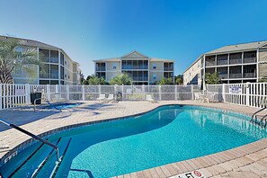 A swimming pool with a white fence, lounge chairs, and a building with balconies in the background.