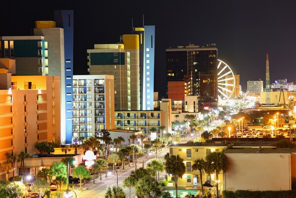 A cityscape at night with modern buildings, a prominent Ferris wheel, and a busy street with palm trees.