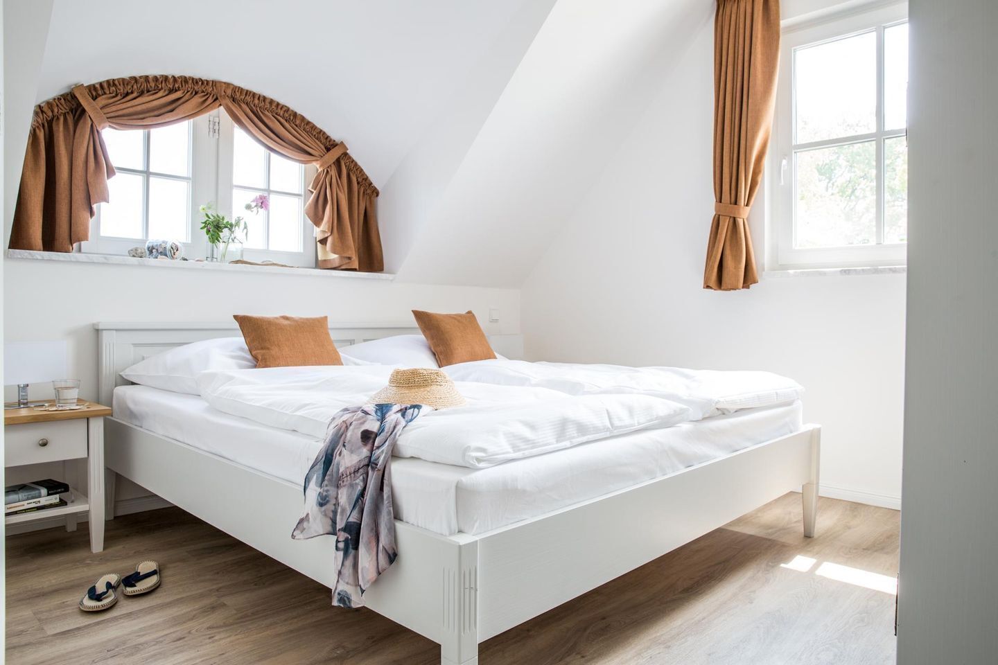 Uma cama bem arrumada com lençóis brancos e duas almofadas castanhas, uma mesa de cabeceira com abajur e uma janela com cortinas.