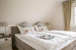 Ein ordentlich bezogenes Bett mit weißer Bettwäsche und dekorativen Kissen, ein Nachttisch mit Lampe sowie ein Fenster mit beigen Vorhängen.