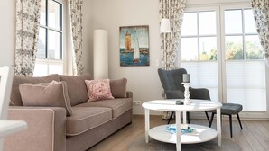 Een woonkamer met een bank, een ronde tafel en een stoel. Er hangt een schilderij aan de muur en een raam met gordijnen.