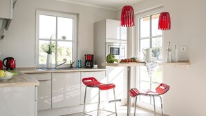 Een moderne keuken met witte kasten, een roestvrijstalen spoelbak, een rode waterkoker en een koffiezetapparaat. Er zijn twee rode barkrukken en een raam met uitzicht op groen.