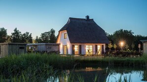 Een huis met rieten dak, een vijver op de voorgrond en een goed verlichte ingang.