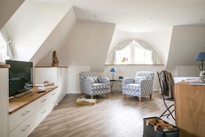 A modern loft bedroom with a sloped ceiling, a large window with curtains, two patterned armchairs, a wooden desk, and a television on a stand.