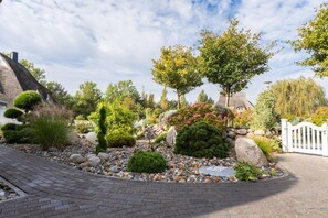 Un jardin aménagé avec différents arbustes, une clôture blanche et une structure surmontée d’un toit de chaume.