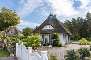Un charmant cottage à toit de chaume, entouré d’une clôture en bois peinte en blanc et d’un jardin aménagé avec diverses plantes et un petit étang.