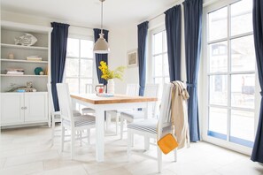 Un comedor luminoso con una mesa de madera, sillas blancas, una lámpara colgante y vistas al exterior a través de amplias ventanas con cortinas azules.