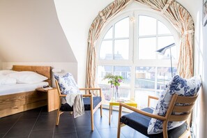 A bedroom with a bed, a wooden nightstand, a chair, a small table, and a large window with curtains.