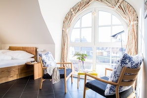 Ein Schlafzimmer mit einem Bett, einem Nachttisch aus Holz, einem Stuhl, einem kleinen Tisch und einem großen Fenster mit Vorhängen.