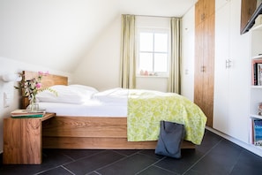 Un dormitorio ordenado con una cama de madera, una mesita de noche con libros, un jarrón con flores y una ventana con cortinas.