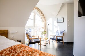 Quarto de hotel com cama, duas cadeiras, uma mesinha e uma janela grande.