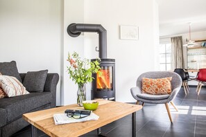 A modern living room with a wood-burning stove, grey sofa, and a dining area with chairs.