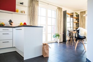 Una cocina moderna con armarios blancos, encimera negra y ventana con cortinas. Hay una bolsa en el suelo y una silla con asiento de pelo.