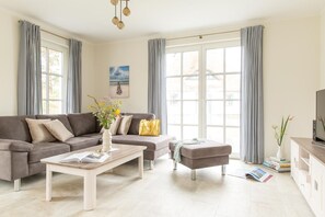 Ein modernes Wohnzimmer mit Ecksofa, Couchtisch, Fernseher und großen Fenstern mit Vorhängen.