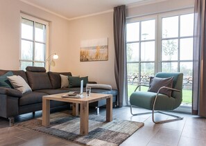 A modern living room with a sofa, a coffee table, and a chair. There is a window with curtains, a painting on the wall, and a lamp.