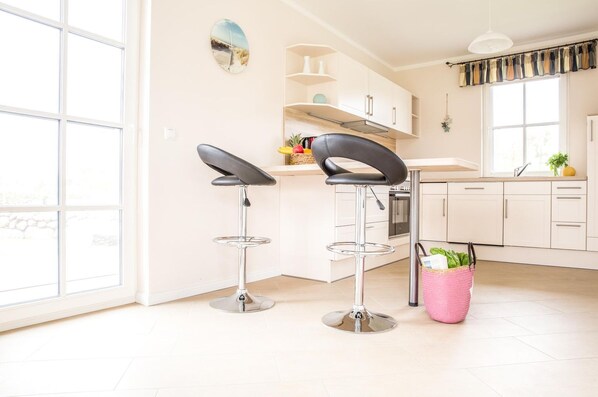 A modern kitchen with two bar stools, a window with curtains, and a basket of vegetables.