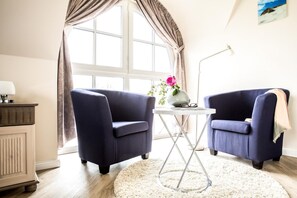 Una habitación con dos sillones azul oscuro, una mesita blanca con un jarrón y vasos, una lámpara y un ventanal con cortinas.