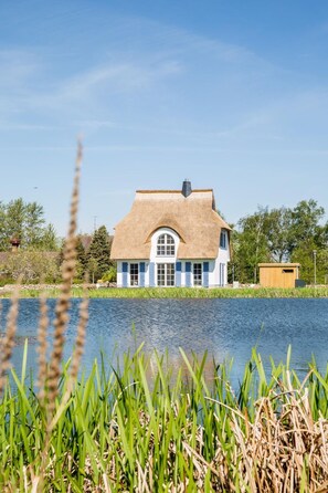 Ein Haus am See mit einem Strohdach und einem kleinen Holzgebäude in der Nähe.