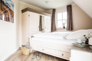 Ein Schlafzimmer mit Bett, einem Kleiderschrank, einem Fenster mit Vorhängen, einem Nachttisch mit Tasse und Blumen sowie einem Korb mit Hausschuhen.