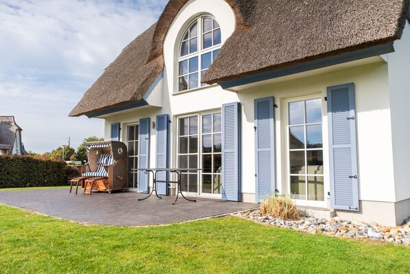 A house with a thatched roof, large windows with shutters, a patio with outdoor furniture, and a well-maintained lawn.