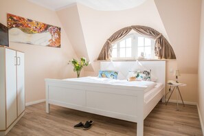 A bedroom with a white bed, a window with curtains, a painting on the wall, and a small table with a lamp.