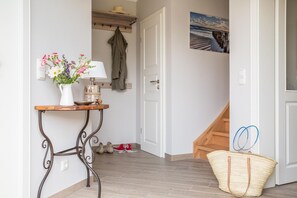 A hallway with a wooden floor, a white door, a small table with a lamp and flowers, and a basket.