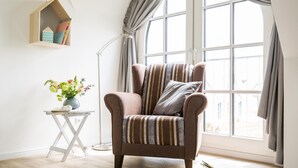 Un sillón cómodo con tapicería a rayas, una mesita auxiliar con un florero y una estantería con libros y una maqueta decorativa de una casa.