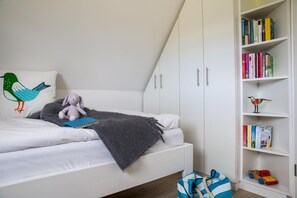Ein Schlafzimmer mit einem weißen Bett, einem Bücherregal mit Büchern und Spielzeug sowie einer grauen Decke auf dem Bett.