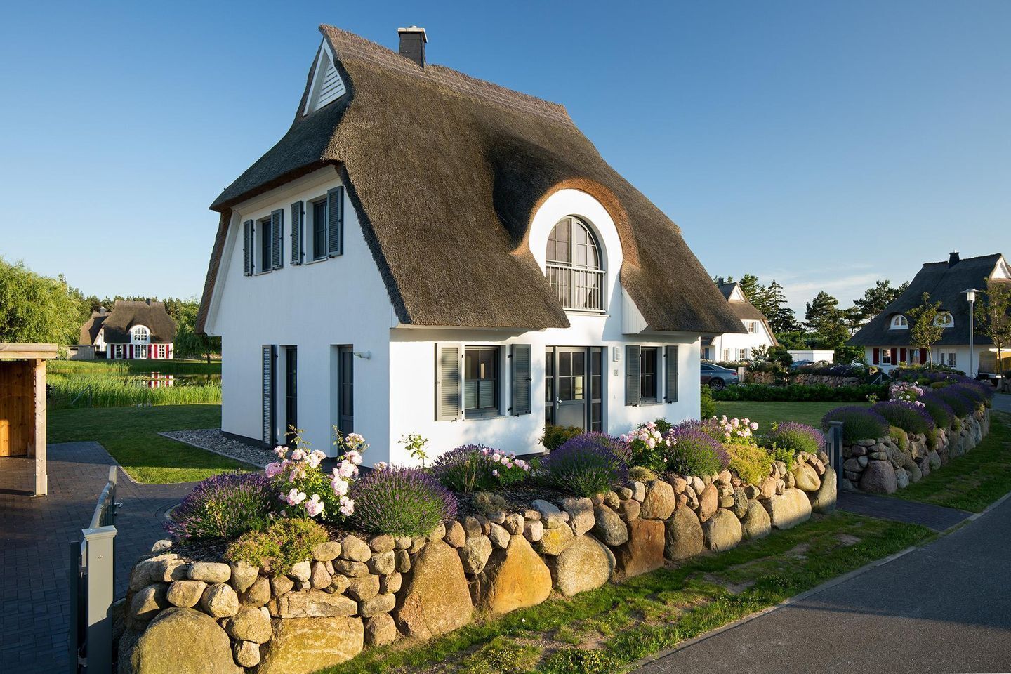 A white house with a thatched roof, a stone wall, and a garden with flowers.