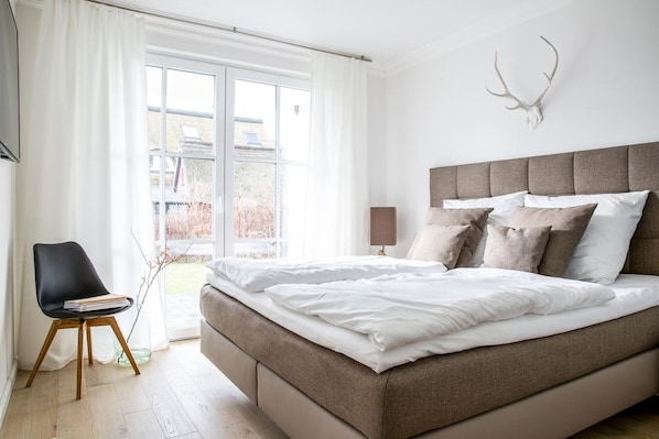 A bedroom with a bed, a chair, a lamp, and a deer skull on the wall.