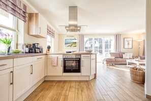 A modern kitchen with a built-in oven, a dining area with a sofa, and a large window providing natural light. - Haubenlerche 20 Vacation Home - Haubenlerche 20 Vacation Home - Close to Nature (Fuhlendorf)