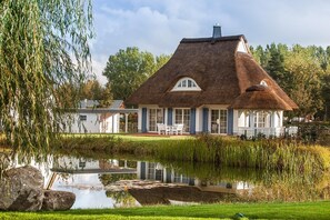 A thatched-roof house with a pond in front, surrounded by greenery.