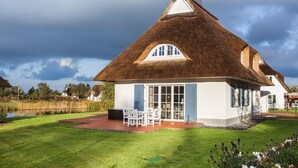A thatched-roof house with a white exterior, a small patio with outdoor furniture, and a pond in the background.