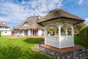 A gazebo with a thatched roof, surrounded by a well-maintained lawn and a house with a similar roof style. - Haubenlerche 20 Vacation Home - Haubenlerche 20 Vacation Home - Close to Nature (Fuhlendorf)