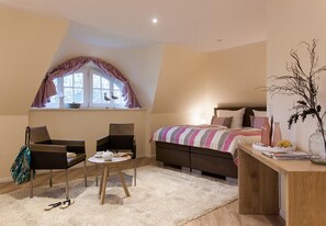 Ein Schlafzimmer mit Bett, Schreibtisch, Stuhl, Fenster mit Vorhängen und einer Vase mit Ästen.