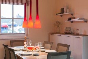 A dining area with a table set for four, a bottle of wine, and a bowl of watermelon. There are three pendant lights above the table, and a window with blinds to the left.