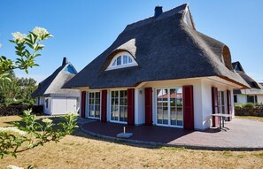 A thatched-roof house with a white exterior and red shutters, featuring a large glass door and a small patio area.