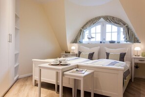 Ein Schlafzimmer mit schrägen Wänden, einem Bett mit gestreifter Bettdecke, Nachttischen mit Lampen und einem Fenster mit Vorhängen.