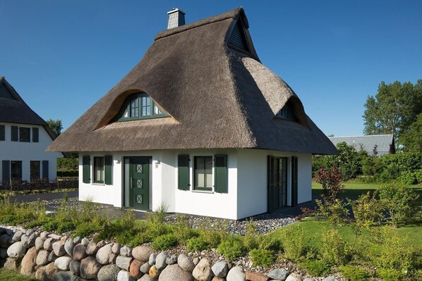 A thatched-roof cottage with a green door and shutters, surrounded by a stone wall and greenery.