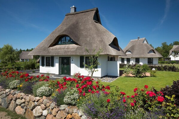 A traditional thatched-roof house with a white exterior, surrounded by a well-maintained garden with colorful flowers and a stone wall.
