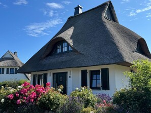Une maison traditionnelle à toit de chaume, dotée d’une façade blanche, de nombreuses fenêtres et d’un jardin aux fleurs multicolores.