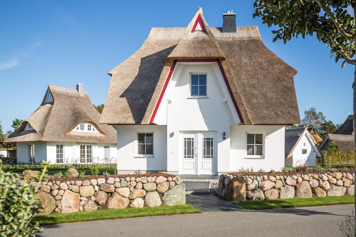 Ein traditionelles Haus mit einem Strohdach, heller Fassade, einer mittleren Tür und einer Steinmauer davor.
