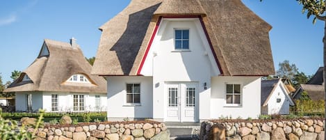 Ein traditionelles Haus mit einem Strohdach, heller Fassade, einer mittleren Tür und einer Steinmauer davor.