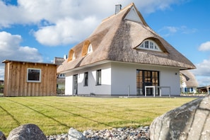 Una casa de techo de paja con fachada blanca, una pequeña construcción de madera a la izquierda y un césped cuidado frente a ella.