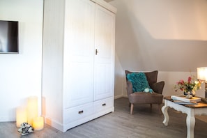 Una habitación con un armario blanco grande, un sillón marrón con un cojín verde azulado, una mesita blanca pequeña con una lámpara y un libro.